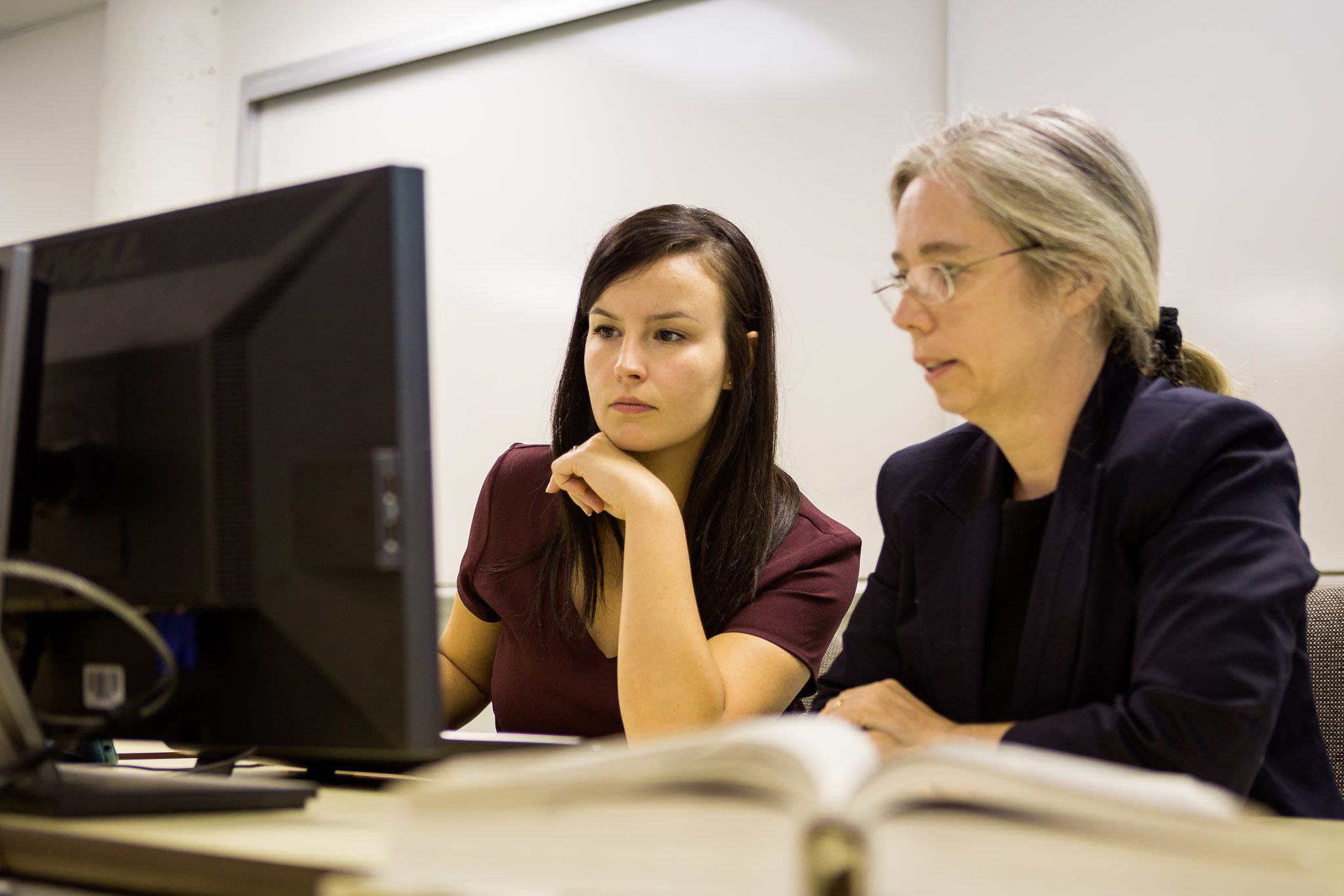 Dans une salle de classe blanche, deux individus d’apparence féminine analysent ensemble le contenu affiché à l’écran d’un ordinateur. Photo.