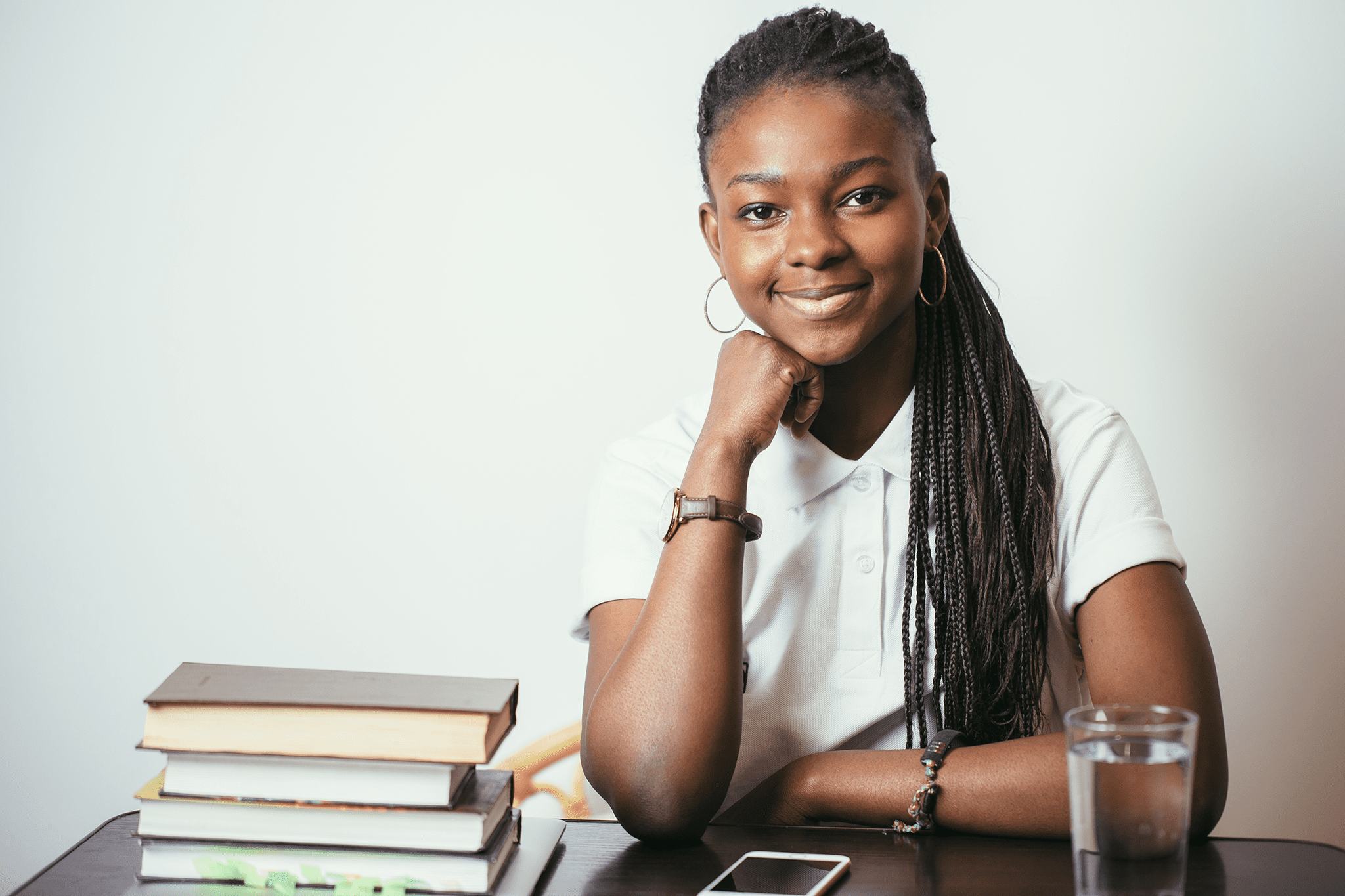 Une jeune fille noire qui semble confiante est accoudé sur un bureau et est entourée de livres, crayons et d'un verre d’eau. Elle est prête à se mettre au travail. Photo.