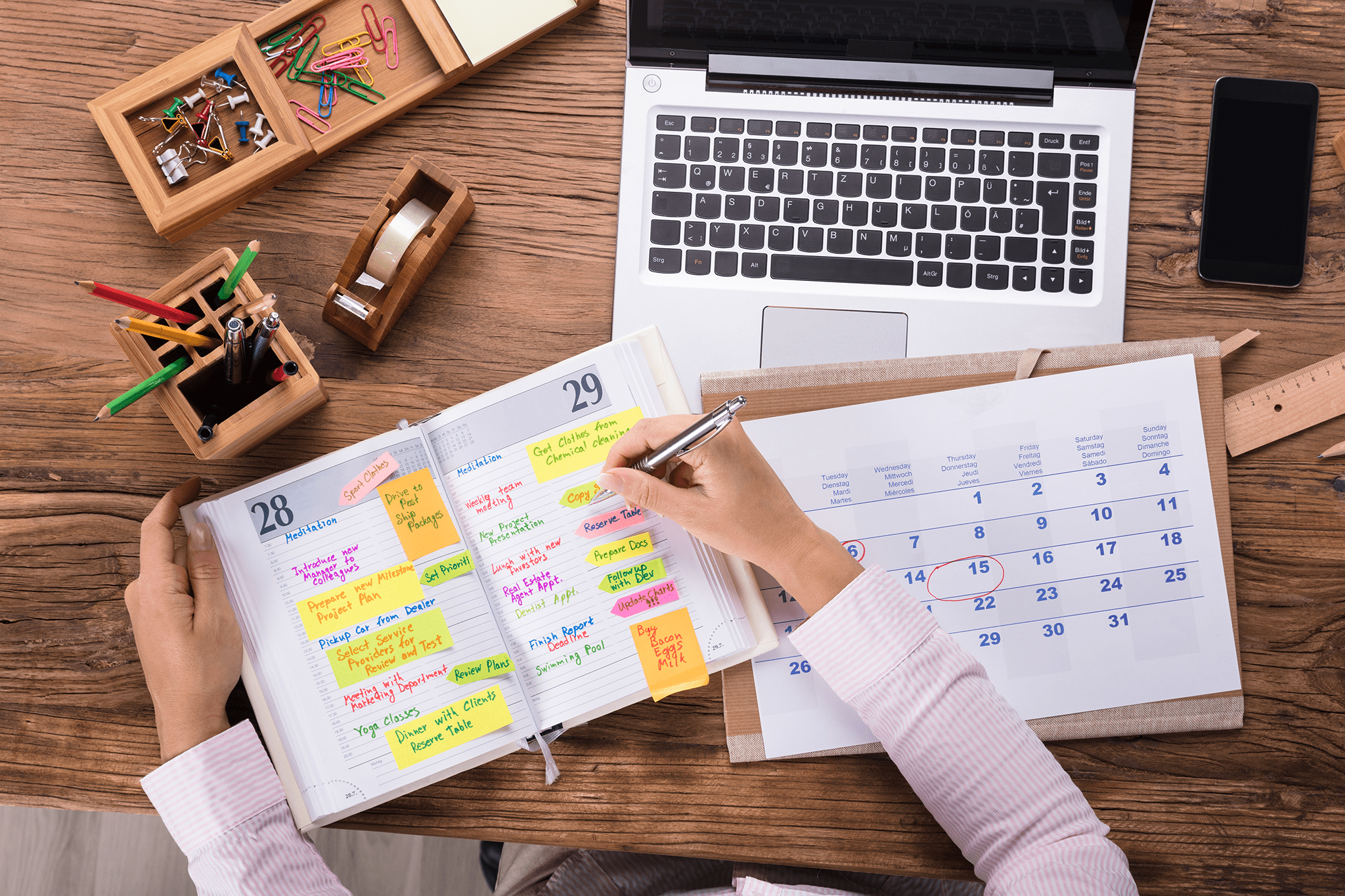 Une personne prend des notes dans son agenda qui est structuré avec du surlignage en couleur et des post-it. Elle à côté d’elle un agenda un ordinateur portable une roulette de ruban gommé, un porte-crayon ainsi qu’un rangement pour ses trombones. Photo.