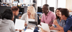 Cinq personnes adultes d’âges différents, discutent autour d’une table. Ils ont en main des documents papier et des tablettes devant eux. Photo.